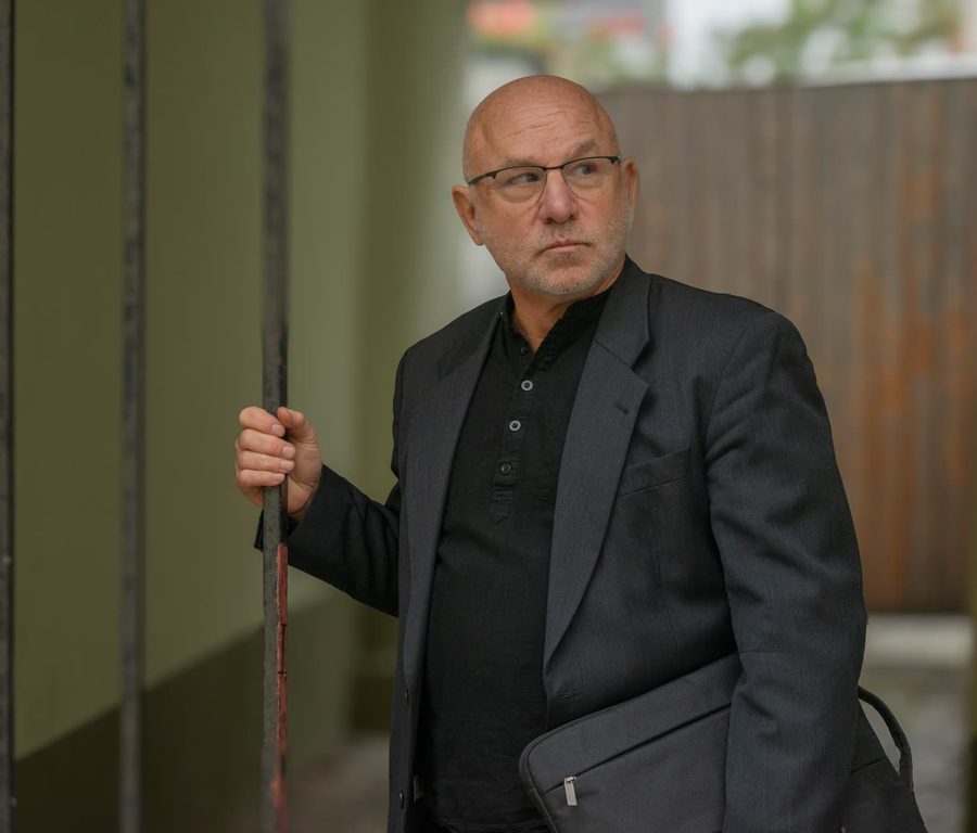 Rechtsanwalt Dr. Donat Ebert in dunklem Anzug mit Laptop-Tasche, an einem Gitterstab lehnend